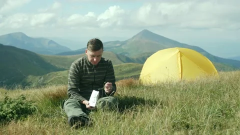 A man on a trip in the mountains sits and eats. A tourist has lunch near a tent Stock Footage 214323634