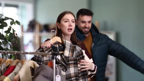 Man try on down jacket, woman elect plaid coat. Family mutual shopping time Stock Footage 330043710