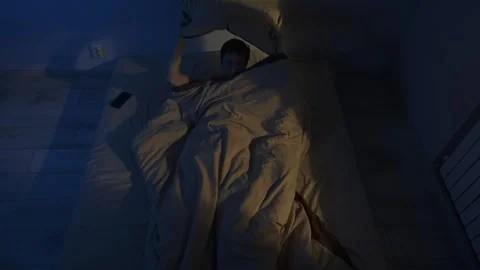Man turning in bed unable to sleep, warm yellow streetlight through window Video stock 321710085