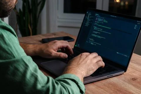 Man types on laptop in dimly lit room while receiving scam message on phone Photos
