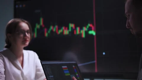 Man types on a laptop in front of a large screen with stock charts in an Stock Footage 317167877