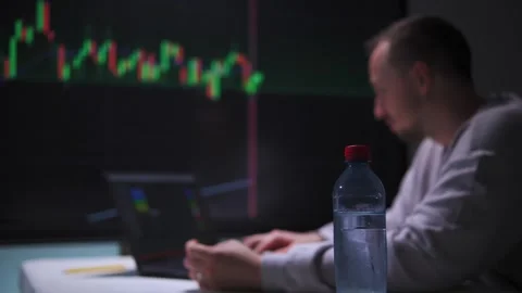 Man types on a laptop in front of a large screen with stock charts in an Stock Footage 317170757