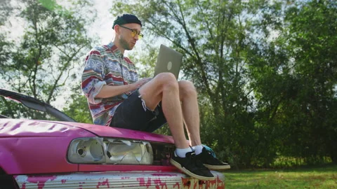 Man types on laptop keyboard sitting on car hood outside Stock Footage 233900927