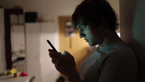 Man types a message with tense expression while leaning against a wall in dim ev Stock Footage 313842882