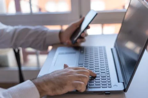 Man typing on a laptop and checking the mobile phone while teleworking. Stock Photos