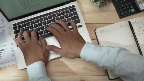 Man typing on a laptop and writing notes in a notebook at a desk Stock Footage 346771320