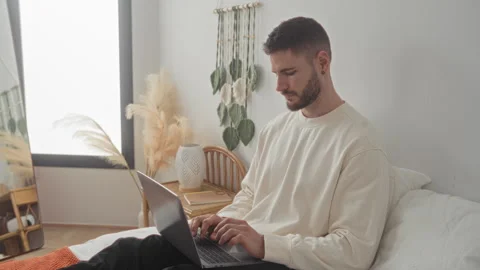 Man typing on laptop on bed in bedroom with white bedding, rattan bedside t.. Stock Footage 337362277