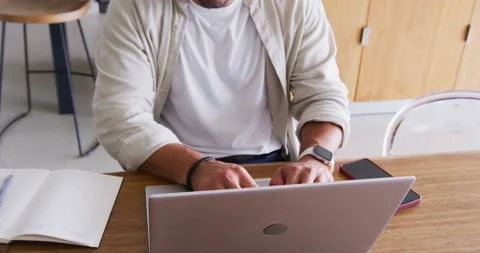 Man typing on laptop at kitchen table, wearing beige shirt, white headphones, Stock Footage 341320838