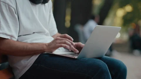 Man typing on laptop outdoors while seated wearing headphones 库存影片 340859080