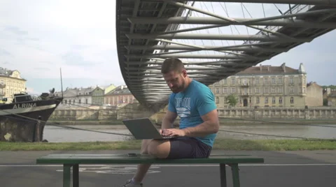 Man is typing on the laptop, sitting on bench against the river. Vídeos de archivo 65188077