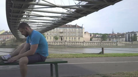 Man is typing on the laptop, sitting on bench against the river, pan shot. Vídeos de archivo 65205143