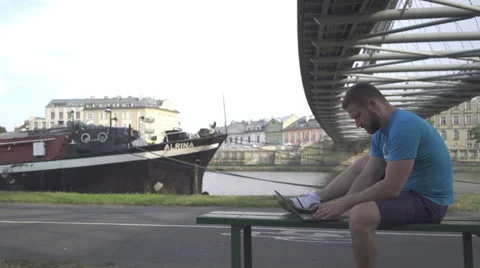 Man is typing on the laptop, sitting on bench against the river, pan shot. Vidéo 65205194
