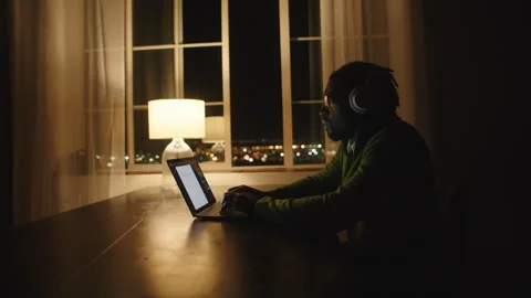 Man typing on laptop while sitting at table under lamp at night Stock Footage 312584283