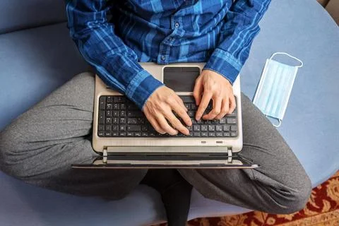 A man is typing on a laptop while sitting on the couch. The medical mask is n Stock Photos