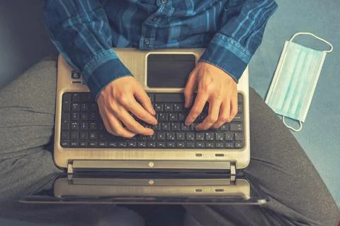 A man is typing on a laptop while sitting on the couch. The medical mask is n Stock Photos