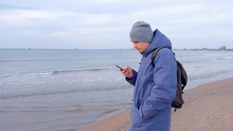 Man is typing message on smartphone in coastal town at sea in winter, side view. Stock Footage 117903972