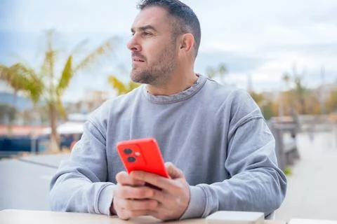 Man typing on red smartphone while sitting outdoors near palm trees and port Stock Photos