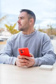 Man typing on red smartphone while sitting outdoors near palm trees and port Stock Photos
