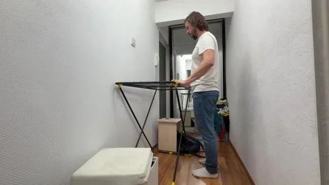 Man unfolding a clothes drying rack in a narrow hallway at home, preparing space Stock Footage 335645727