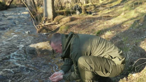 A man in uniform washes his face with water from a forest stream. Видео 194950519