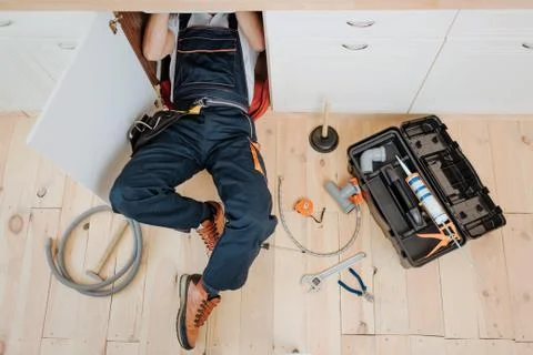 Man in uniform work in kitchen under sink. He repair water leaking. Hose and 库存照片