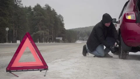 A man urgently changes a wheel on a car in winter. A bright red triangle on a Stock Footage 144484366
