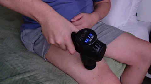 Man uses battery-powered percussion massage gun on his right thigh Stock Footage 331124756