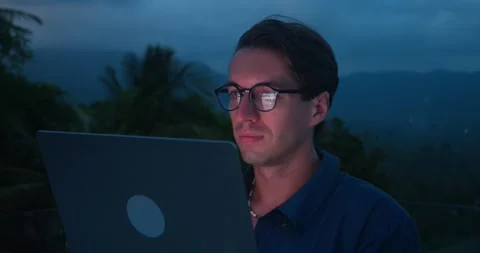 Man uses laptop outside in evening. Light from laptop screen illuminates man's Stock Footage 260108710