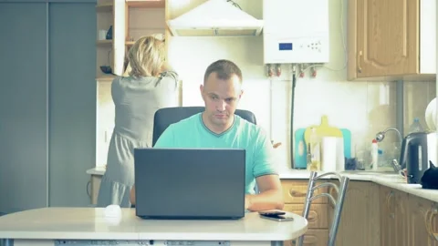 Man uses laptop at the table at home in the kitchen, woman prepares food Stock Footage 138273743