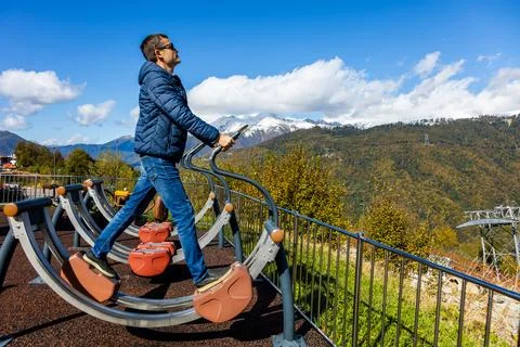 Man uses outdoor exercise machine on mountain with snow-capped peaks Stock Photos