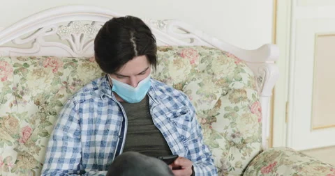 Man uses phone when resting on sofa, lowers mask and is weary on isolation Vídeo Stock 130434586