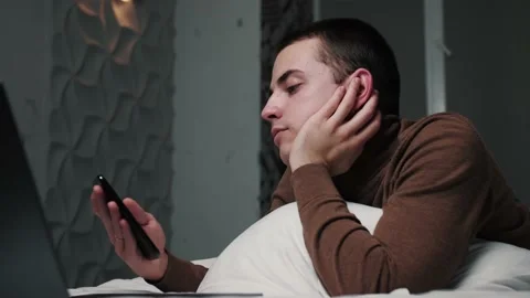 Man uses a phone while lying on bed. Handsome young concentrated caucasian man Stock Footage 144311377