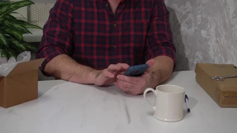 A man uses a phone while sitting at a table with parcels. The guy taps on the 库存影片 305025462