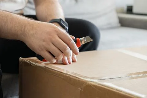 A man uses a sharp utility knife to cut adhesive tape on a brown cardboard box, Stock Photos