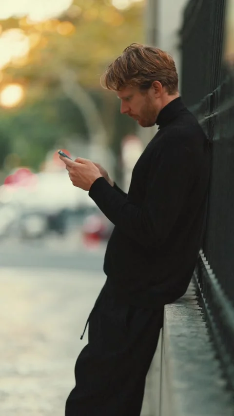 Man uses smartphone while leaning against a wall in an outdoor setting during Stock Footage 346702436