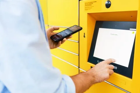 Man uses a smartphone while tapping the touchscreen of a yellow parcel lock.. Foto stock