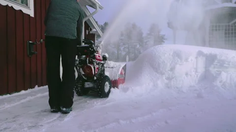 A man uses a snow blower to clear thick snow near a red wooden house during.. Stock Footage 287073714