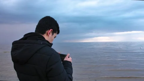 A man uses a tablet to capture the serene sea despite the cloudy sky Stock Footage 281504100