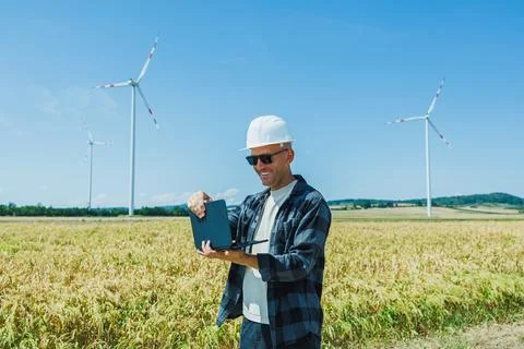 Man uses a tablet to check data in a field near wind turbines on a sunny day 스톡 사진