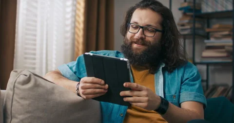 A man uses a tablet on the couch, talks to parents via video camera, waves hello 動画素材 218202863