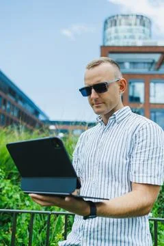 Man uses a tablet outdoors while standing by green plants near a modern bui.. Stock Photos