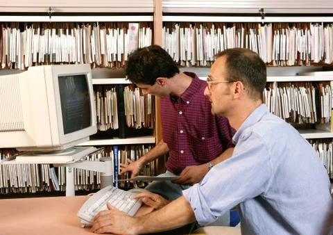 Man using computer, other man looking at files on shelves Stock Photos