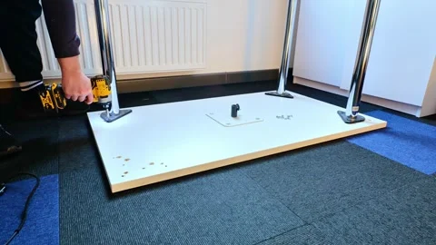 Man Using Electric Drill to Assemble Table Legs Indoors on Carpeted Office Floor 스톡 동영상 306767477