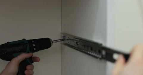 Man using a electric drill to attach a metal drawer slide to a piece of Видео 329763073
