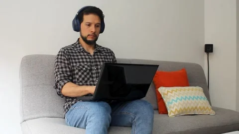 A man using a headset sitting on a grey couch working from home on a laptop Stock Footage 340583327