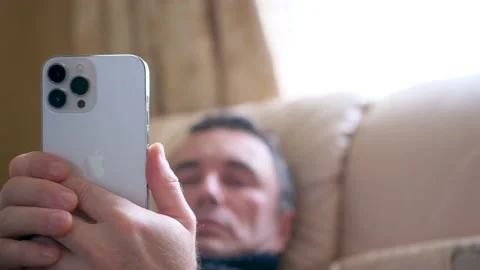Man using Iphone 13 Pro Max against blurred a man lies on the bed resting using Stock Footage 239220910