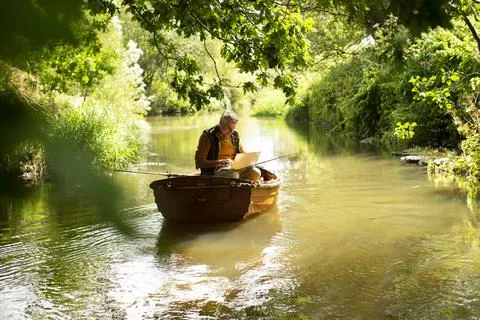 Man using laptop and fly fishing in boat on sunny river 스톡 사진