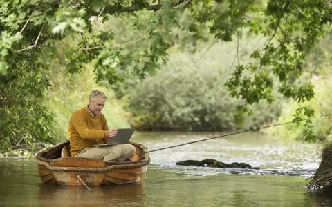 Man using laptop and fly fishing in boat on river Foto stock