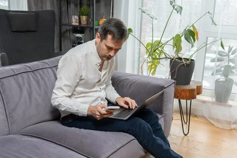 Man using laptop and smartphone while sitting on comfortable sofa at home Stock Photos