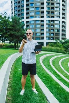 Man using a laptop and smartphone in a grassy area near a modern building o.. 스톡 사진